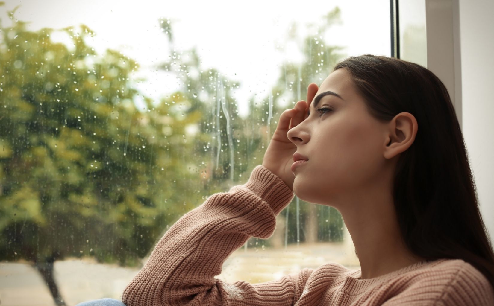 Woman sitting by a rainy window and considering trauma therapy in Massachusetts.